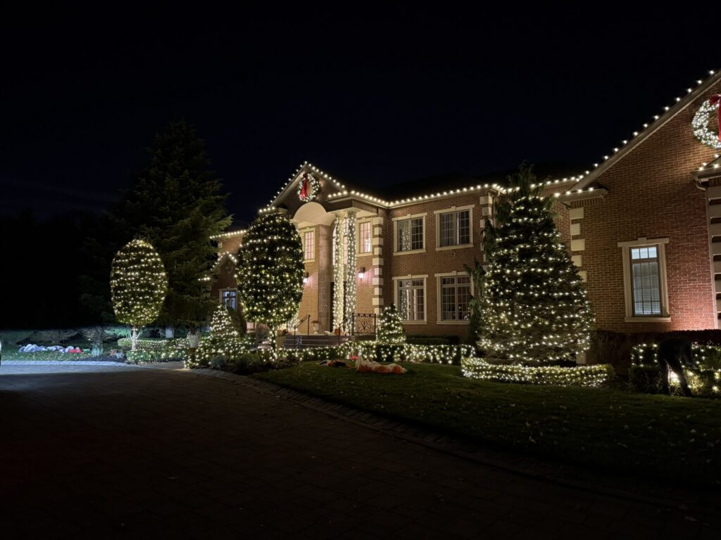 Brick house with a warm white roofline and wrapped evergreens at night