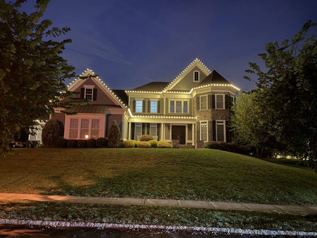 Cottage-style house with a warm white roofline and lit shrubs at dusk