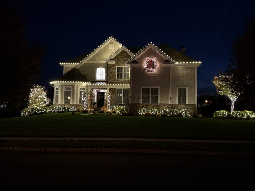 House with a warm white roofline, a wreath and lit shrubs at night