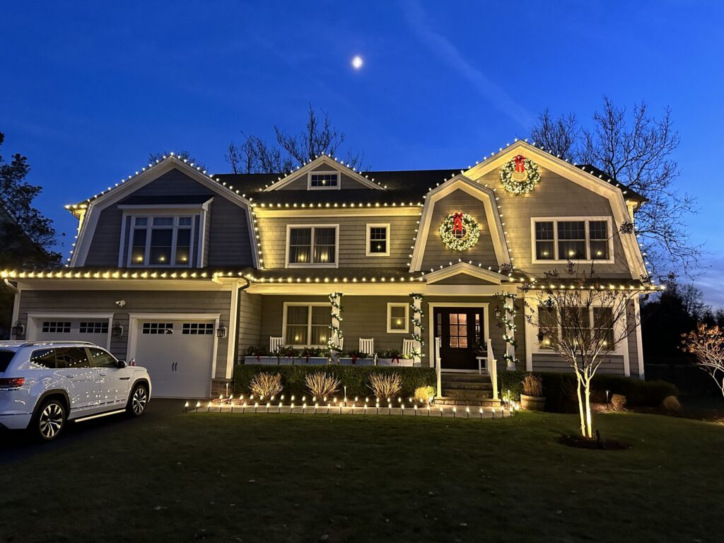 Shingle-style house with a warm white roofline and wreaths at blue hour