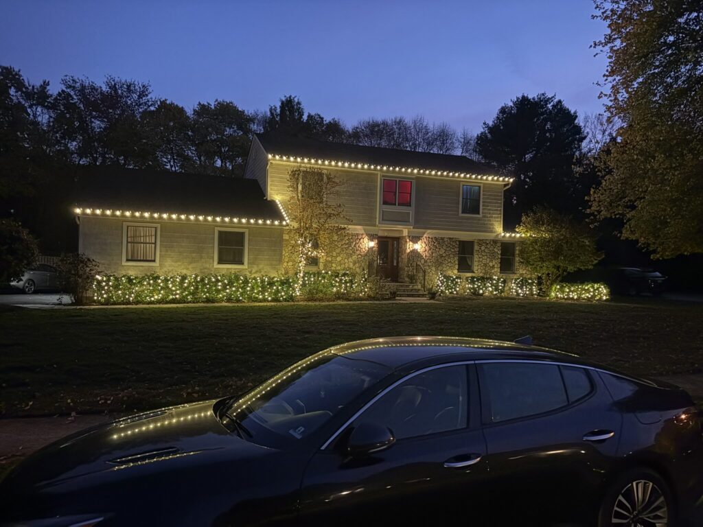 Two-story house with a warm white roofline and lit shrubs at dusk