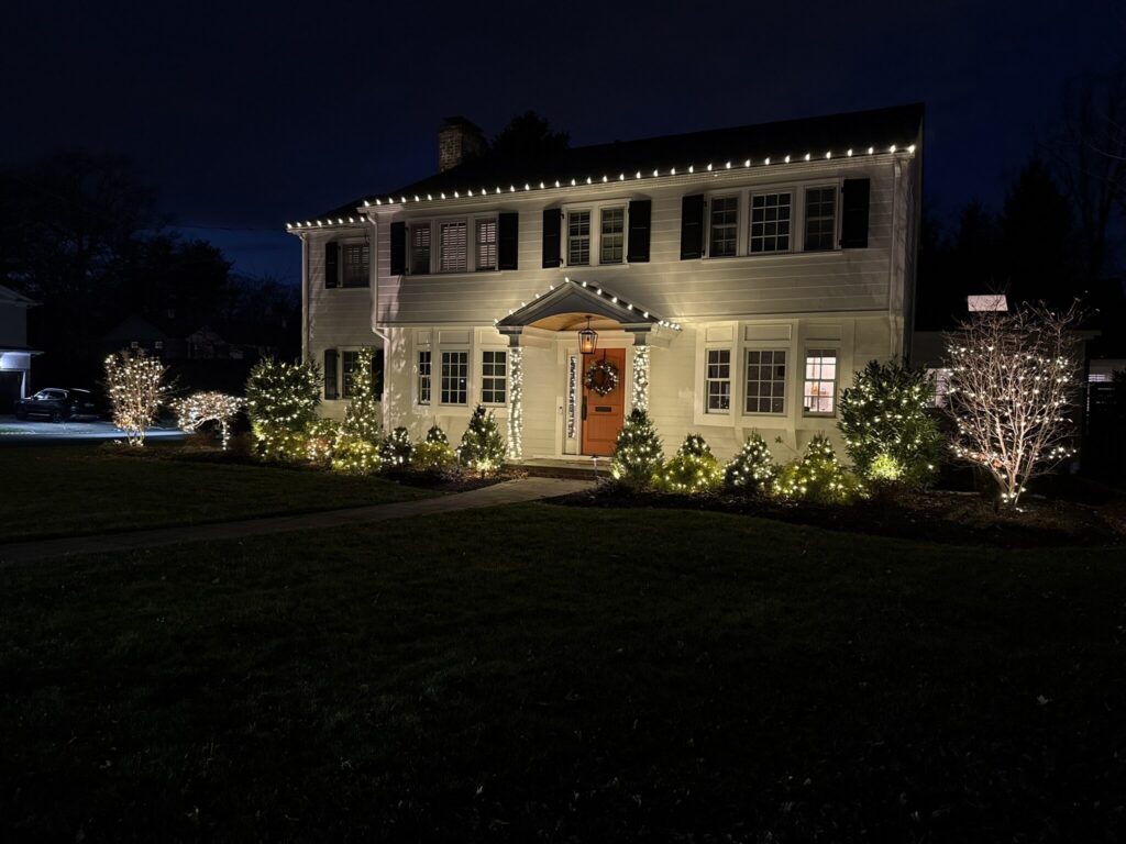 White colonial with a warm white roofline and lit shrubs at night