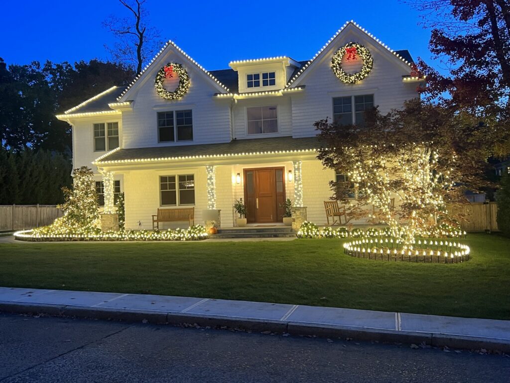 White farmhouse with a warm white roofline, wreaths and wrapped trees at blue hour
