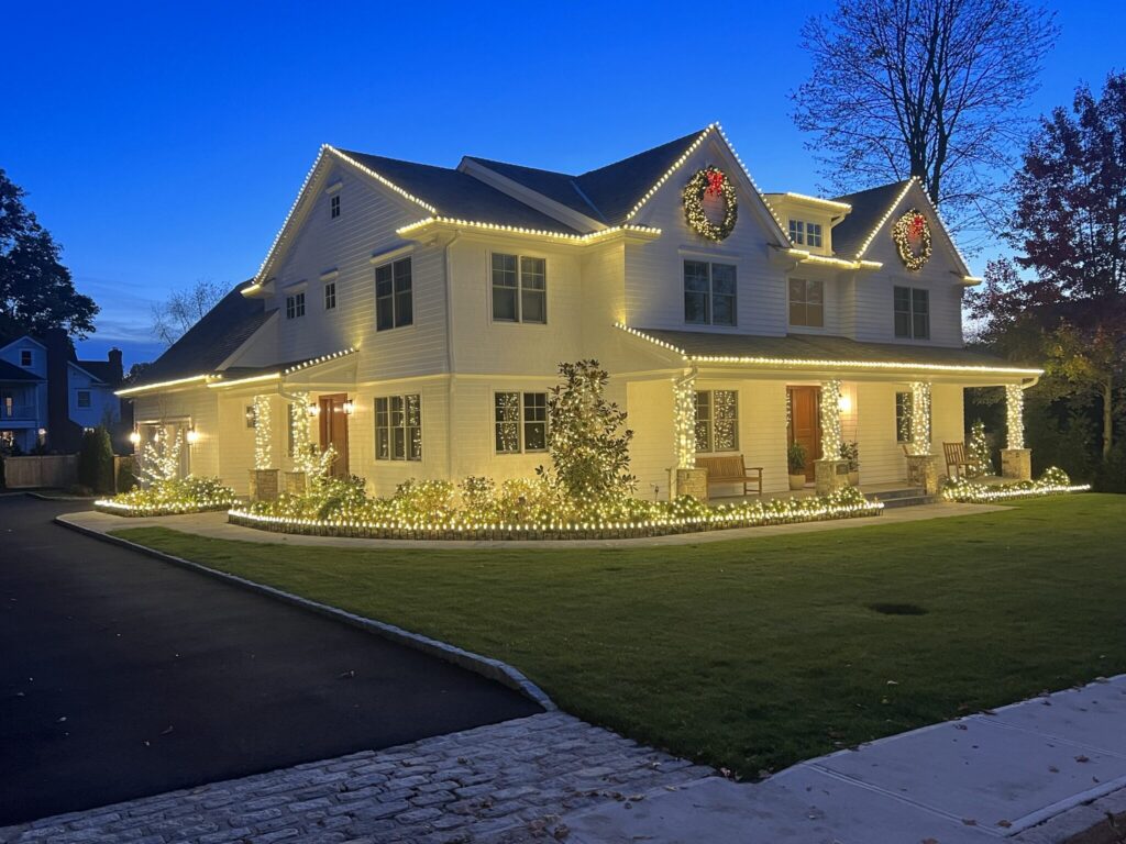 Side view of a white farmhouse with a warm white roofline and wreaths at blue hour