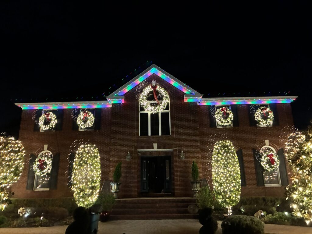 Brick colonial with a permanent roofline in blue and white, lit wreaths and wrapped trees
